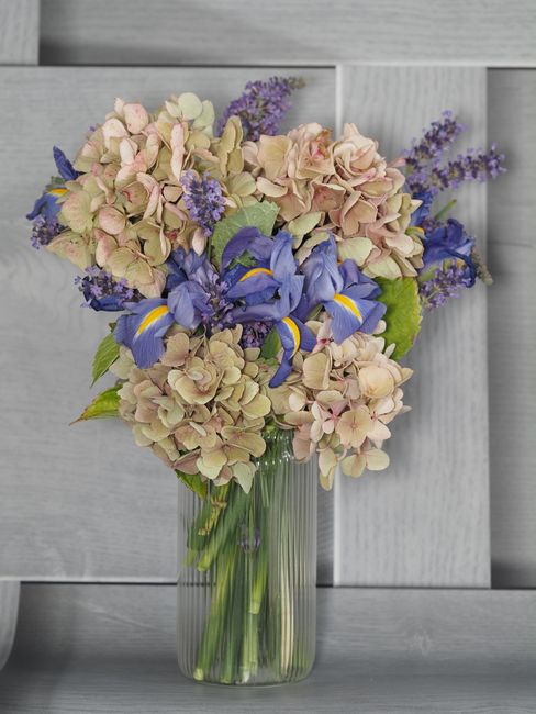 A tall, clear glass vase holding a floral arrangement of pastel pink and cream hydrangeas, vibrant blue iris flowers with yellow accents, and sprigs of purple lavender. The bouquet is arranged in a lo