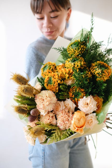 A woman with short brown hair and a light grey top holds a large, vibrant bouquet of fresh flowers. The arrangement features yellow and orange blooms, including roses, carnations, and small clusters o