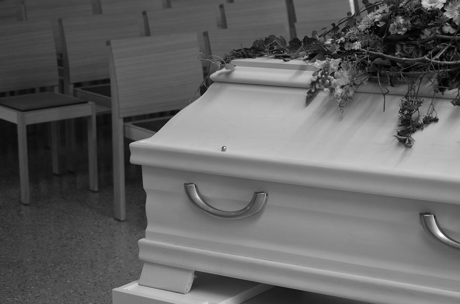 A white coffin with simple metallic handles, topped with a floral arrangement consisting of mixed flowers and greenery, resting on a stand inside a funeral chapel setting. The arrangement includes pur