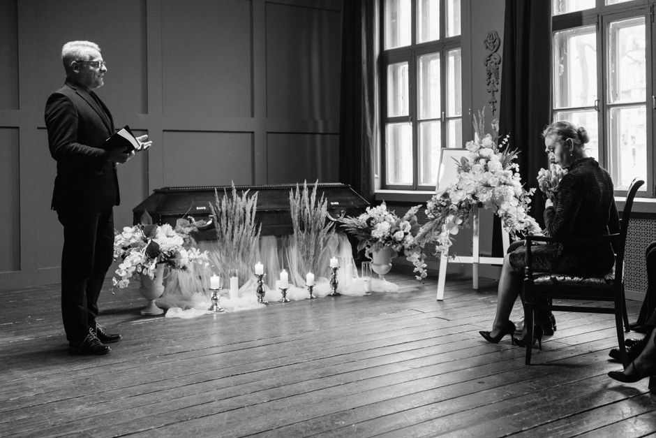 A black-and-white photograph of a floral arrangement at a funeral service inside a chapel. The arrangement features a large, cascading bouquet of fresh white roses, lilies, and other blooms, with deli