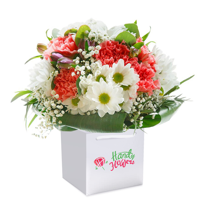 Presented with sophistication and care, this stunning floral bouquet is elegantly positioned in an immaculate white square gift bag, a vision reminiscent of Fortis Green's tranquil charm. A generous mix of coral-pink and red carnations takes center stage, their textured, frilled petals full of tactile richness and energy, evoking the vibrant hues found in local artisanal shops along Fortis Green Road. Interspersed among these are classic white daisies, their striking bright green-yellow centers adding an unmistakable pop of joy, along with ethereal white baby's breath that offers softness and visual lift to the composition. Set toward one side, several delicate buds edged in purple and pink hint at alstroemeria or lily, suggesting life's unfolding promises. A lush collar of broad, deep green leaves provides a natural embrace, grounding the bouquet and enhancing its fullness. The clean white bag, complete with sturdy handles, is designed for graceful transport-whether accompanying a celebration in the heart of the community or a surprise delivery to a friend near Highgate Wood. No extensive branding distracts from the purity of the flowers themselves; rather, attention is drawn to their freshness, impeccable combination, and the intention behind each stem. This composition radiates warmth, elegance, and a sense of occasion-beautifully symbolizing the joy of gift-giving and the closeness found throughout Fortis Green.