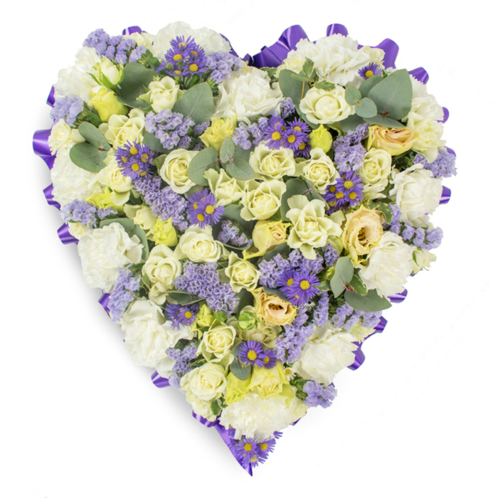 A carefully constructed heart-shaped memorial arrangement set against a plain white studio background, the composition speaking in gentle tones of cream, white and lavender. The outer perimeter is crisply defined by a continuous purple satin ribbon that has been looped and folded to create a neat, formal edge; within this frame the surface is densely filled with cream and pale yellow roses, many held as tight buds for longevity alongside a few softly opening blooms to give depth and life. White carnations are used generously to build volume and a soft, comforting foundation, while tiny pale yellow florets add a whisper of warmth between the larger blooms. Throughout the heart, clusters of lavender-hued statice bring an airy texture, and small violet daisy-like flowers with bright yellow centres provide richer colour notes that draw the eye inward. Rounded, silvery-blue eucalyptus leaves are placed at intervals to give sculptural punctuation and a clean, modern silhouette. Photographed flat on white, the arrangement communicates a quiet dignity and careful craftsmanship - qualities often sought for funeral services, wakes, or graveside tributes in the Fortis Green neighbourhood and nearby Muswell Hill. The overall impression is one of steady compassion and refined simplicity: a sympathetic design that conveys remembrance and respect while remaining elegant and understated.