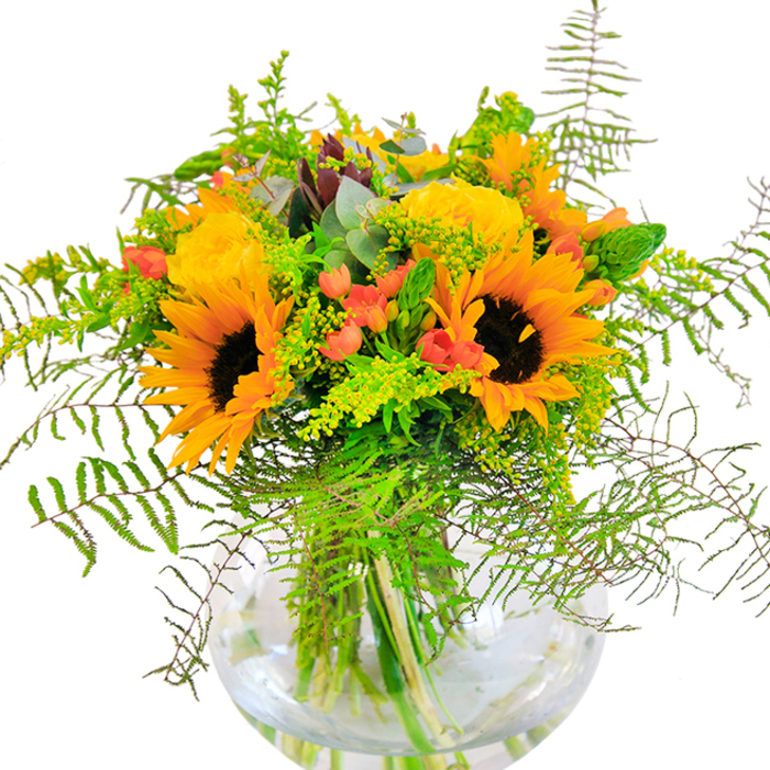 The first sensation from this evocative bouquet is sunlight distilled-an exuberant collection of golden yellows, vibrant oranges, and lush greens that perfectly encapsulates the joyous feel of summer days in Fortis Green. Majestic sunflowers and plush yellow roses bloom side by side, their petals overlapping in warm, generous layers that seem to share the same glowing light as the open spaces of Cherry Tree Wood at midday. Interlaced among these central blooms, glossy red hypericum berries shimmer against the arrangement's soft, velvety petals, their brilliance a playful nod to the colourful market stalls along Fortis Green Road. Mesmerising streams of feathery fern fronds and trailing greenery flow outward, blending seamlessly with subtle, springy goldenrod and thread-like eucalyptus leaves-details that invite touch and reflect the area's garden-rich charm. A clear, bubble-shaped vase holds this abundance, letting the natural beauty of the green stems and pure water shine through, reminiscent of a freshly gathered bouquet from the neighbourhood's own green spaces. Up close, the interplay of textures-from the suede sunflowers to the delicate garden rose spirals-comes alive, accompanied by a gentle, clean fragrance that stirs thoughts of nearby parks and sunlit gardens. Whether chosen to celebrate a joyful birthday, an anniversary, or to mark any special occasion with cheer, this arrangement is a heartfelt tribute to the vibrant community and seasonal splendour of Fortis Green.