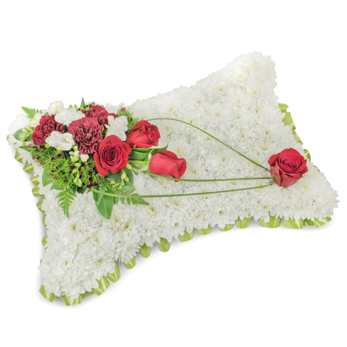 An elegantly sorrowful floral pillow is shown in soft lighting, its rectangular silhouette formed entirely from compact, snowy white chrysanthemums that create a plush, tactile surface meant to evoke peace. The tightly arranged blooms catch a subdued light that highlights the roundness of each petal cluster and the cushion's slight curvature. Around the perimeter a whisper of pale green ribbon or leaf peeks out, lending a delicate frame and a suggestion of new growth. On the upper left a lush cluster of deep red roses and dark-hued carnations sets a warm, dignified tone; the roses vary from tight buds to open cups, their satin petals rich against the white background. Interspersed tiny white florets - likely freesias or similar spray blooms - add airiness and a soft, clean scent, while feathery fern fronds and broader glossy foliage provide a verdant underlay. From this primary group a trio of slender green bear-grass arcs flows diagonally across the chrysanthemum field toward the bottom right, their elegant curves guiding the gaze to a single, flawless red rose that appears to rest gently at the pillow's corner. The arrangement reads as a classical expression of tribute and remembrance, measured and sincere; it is the kind of piece many residents request for funerals and memorial services in the Highgate and Fortis Green neighbourhoods, where understated sympathy tributes are preferred for services at local churches and funeral homes.