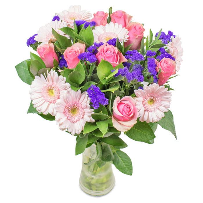 An enchanting floral arrangement crafted near the heart of Fortis Green, this bouquet is artfully presented in a clear glass vase and set against a serene, light-infused backdrop. At its core, roughly eight pink roses, each in a different stage of bloom - some tightly furled, others just beginning to reveal their spiral petals - exude classic charm and intimate romance reminiscent of gatherings along Fortis Green Road. Surrounding these, five or six delicate gerbera daisies display their soft, round faces with pastel pinks and almost white centres, infusing the bouquet with a gentle tenderness. Vibrant clusters of purple statice are interwoven throughout, their bold, velvety blossoms lending both drama and texture and instilling the piece with lively contrast, much like the artistic vibrancy found nearby at Alexandra Palace. Deep emerald foliage forms a lush, abundant base, supporting the blooms and imparting a breath of freshness reminiscent of local woods and greenways. The arrangement's stems are visible through the water-filled vase, enhancing the composition's natural character and celebrating the joy of fresh flower delivery to homes and offices across Fortis Green. Suitable for a range of occasions from heartfelt birthdays and Mother's Day tributes, to anniversaries celebrated in sun-dappled living rooms, this bouquet conveys an unmistakable sense of affection, celebration, and everyday beauty drawn from the rich tapestry of its local surroundings.