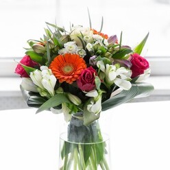 This breathtaking bouquet, hand-crafted in the heart of Fortis Green, is an elegant interplay of vibrant hues and luxurious textures, capturing the beauty of local gardens in one flawless arrangement. At its heart, lush gerbera daisies glow-two in deep orange, recalling the fiery sunset seen from atop Muswell Hill, and two softer, creamy peach blooms with velvety brown centres, exuding a gentle embrace. Interlaced are rich fuchsia roses, tightly furled and opulent, their depth echoing the intimacy of nearby mews and hidden courtyards. Delicate clusters of pristine white alstroemeria and petite spray roses add a gentle radiance, like a scattering of light across Fortis Green's leafy avenues at dawn. Gathering around the ensemble, glossy, deep green leaves-some large and paddle-shaped, others slender and pointed-are carefully sculpted to create a protective collar, much like the canopy of Highgate Wood. Gentle touches of lighter pink blossoms and dainty white fillers lend extra depth, forming an abundant, rounded shape brimming with optimism. The clear glass vase showcases the bouquet's verdant stems, basking in the ambient daylight poured through nearby windows, contributing to its organic vibrancy. Light falls softly across the scene, enhancing the natural nuances of every petal and leaf. This striking display would feel perfectly at home brightening a Fortis Green dining table, welcoming guests in a local café, or marking a milestone with understated grace. Every element sings with care and expertise, presenting a premium expression of nature's beauty rooted in the local community.
